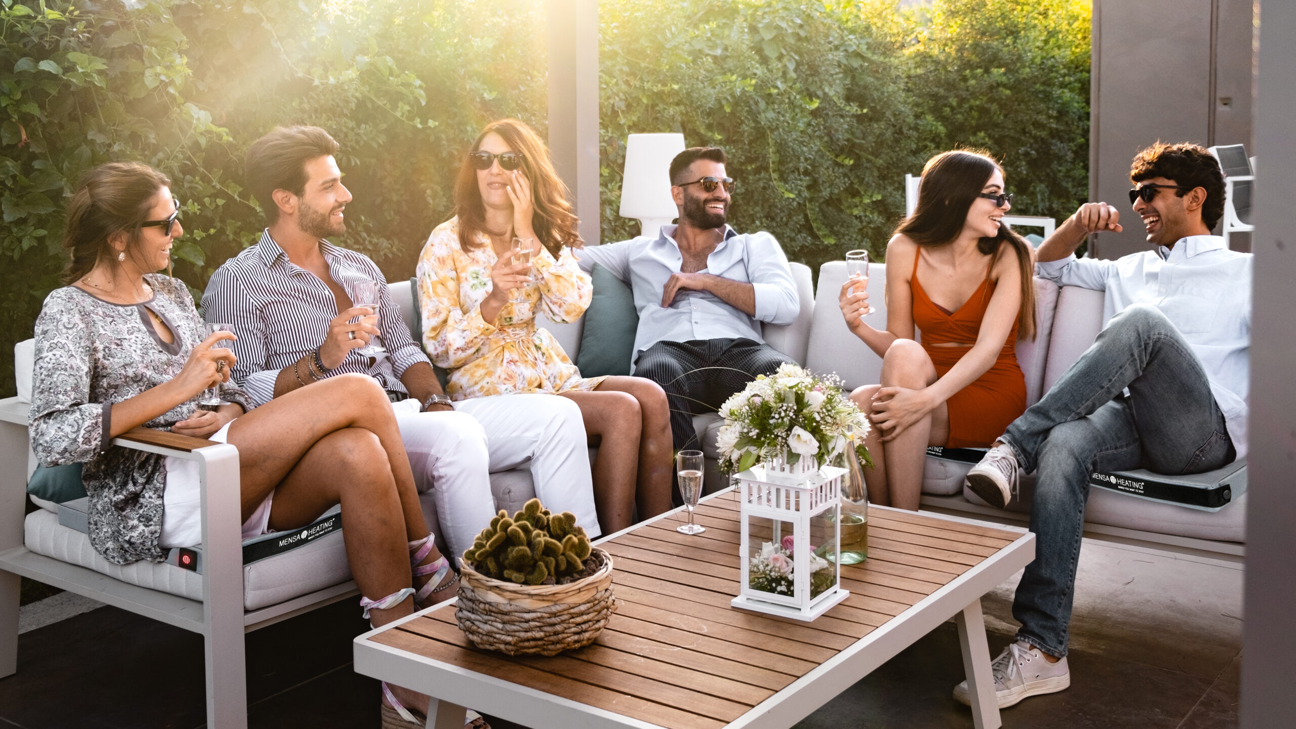 Group of cheerful young people carefree sitting together drinking wine at luxury garden - friends get together sitting on a couch in the terrace backyard having fun drinking champagne