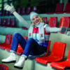 Beautiful blond young woman in winter clothes and  white skates sitting on red tribune. Girl is ready for skating on ice rink.