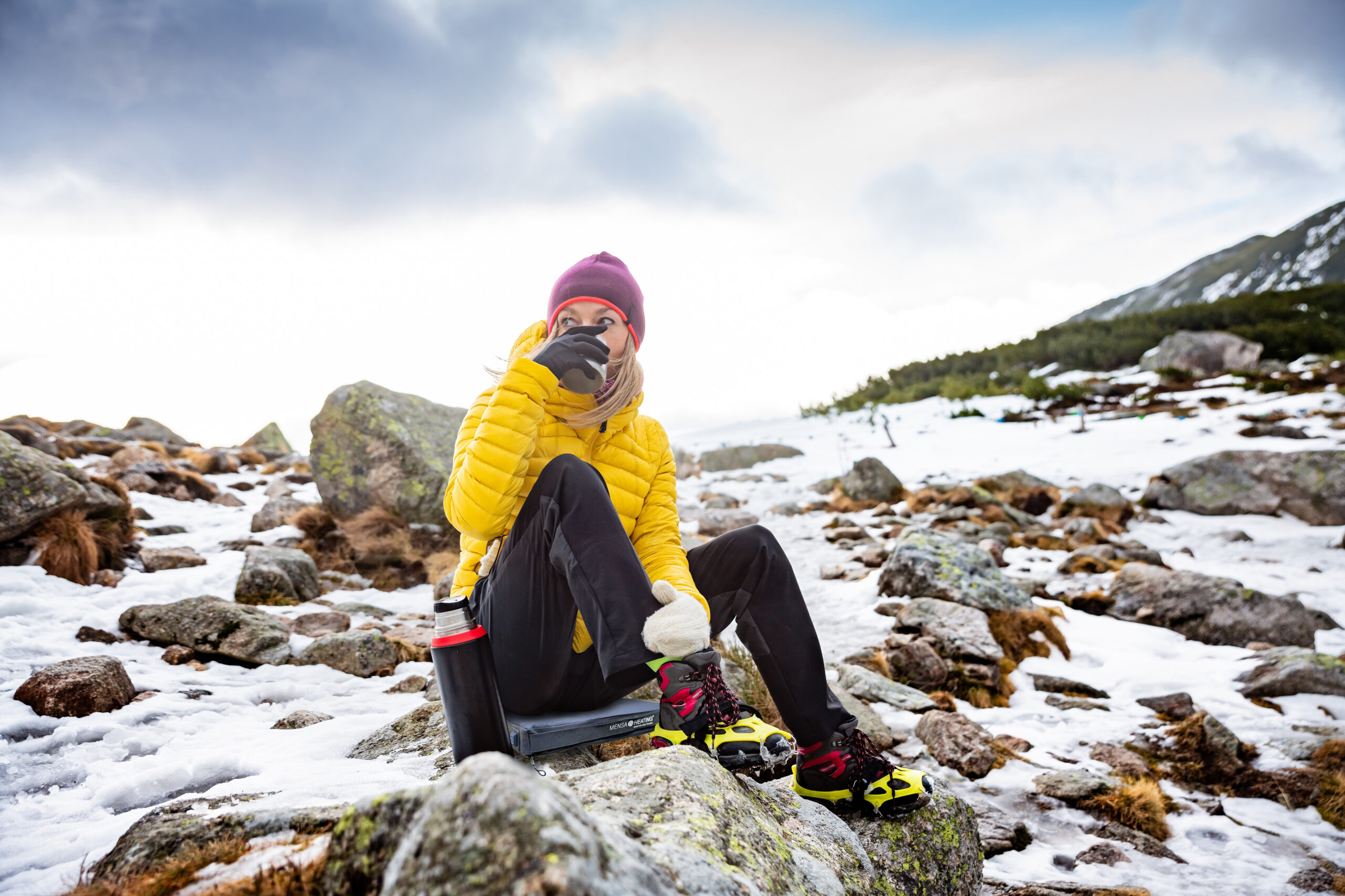 A woman traveler in a yellow down jacket is resting and drinking hot tea on a cold, windy day.